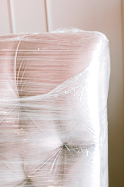 A large piece of furniture, possibly a sofa or mattress, wrapped securely in clear plastic wrapping to protect it during moving. The furniture is positioned inside a home near a doorway or passageway,