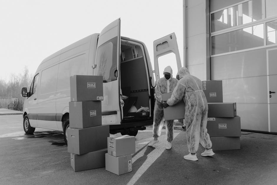 Two removal specialists dressed in protective overalls, face masks, and hair covers work together to load a cardboard box into the rear of a large white moving van parked outside a modern building wit