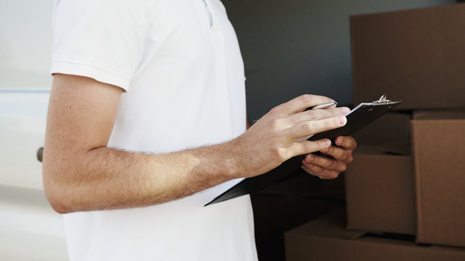 Two removal specialists dressed in protective overalls, face masks, and hair covers work together to load a cardboard box into the rear of a large white moving van parked outside a modern building wit