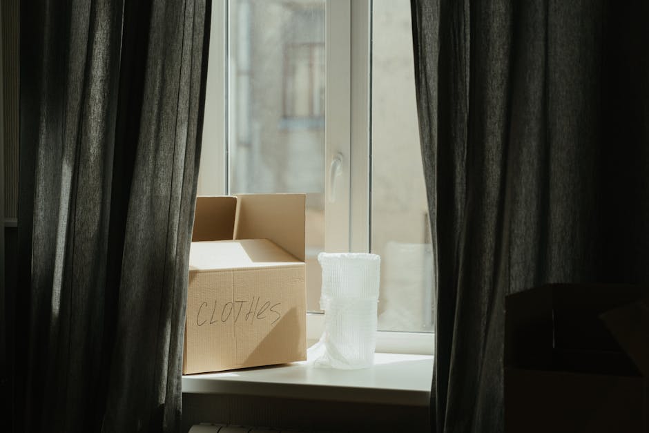 A cardboard box labeled 'CLOTHES' is positioned on a windowsill inside a room, with the box partially open revealing some clothing items inside. Next to the box, a white fabric or plastic-wrapped bund