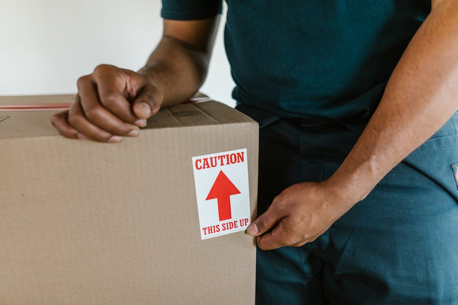 A cardboard box labeled 'CLOTHES' is positioned on a windowsill inside a room, with the box partially open revealing some clothing items inside. Next to the box, a white fabric or plastic-wrapped bund