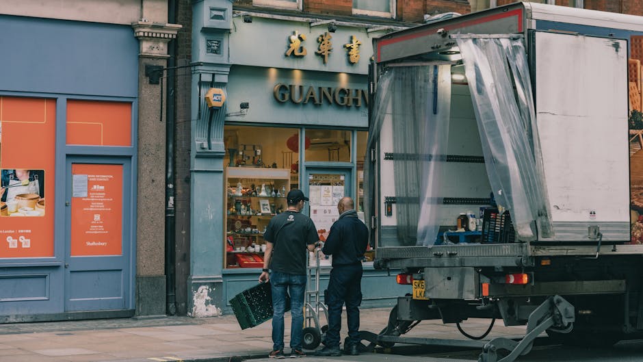 Two men sitting on the floor inside the rear of a moving van during a house relocation, surrounded by packed cardboard boxes and protective foam assists. One man is wearing a beige beanie, a blue and